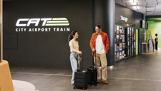 Mann und Frau mit Koffern stehen lächelnd im Warteraum des Wiener City Airport Trains CAT