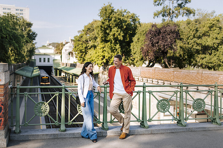 Mann und Frau lehnen lächelnd an Geländer mit Blick auf eine U-Bahn Station und Gleise